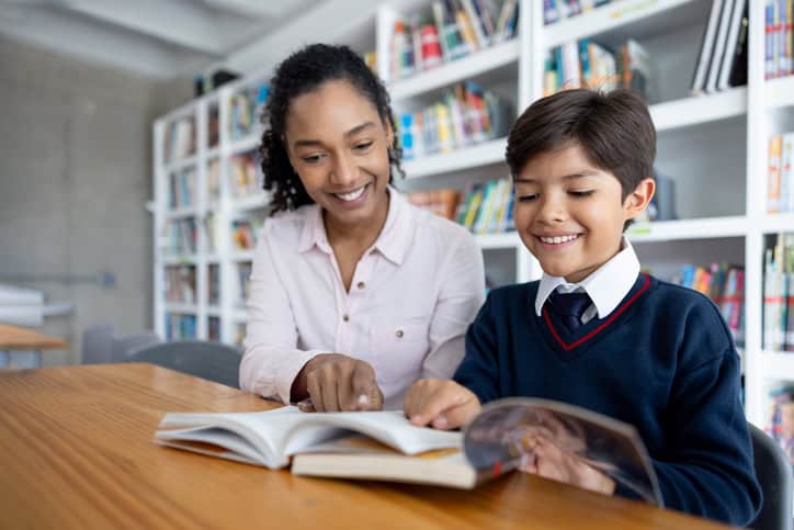Happy teacher helping an elementary student while reading a book at the library - education concepts